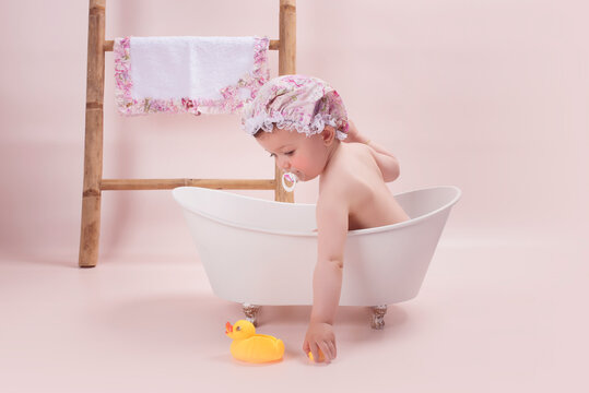 Ni&ntilde;a con chupete que se da un ba&ntilde;o en una mini ba&ntilde;era, lleva un gorro de ducha y una toalla a conjunto colgada en una escalera de madera