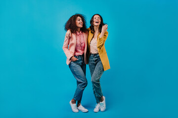 Two joyful girls posing on blue studio background. Full length photoshoot of stylish female models.