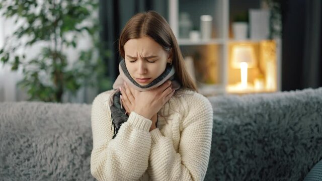 Sad young woman in warm sweater and scarf sitting suffering from sore throat while staying at home. Concept of sick and tired.