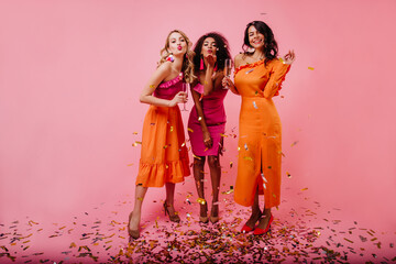 Satisfied girls standing on confetti. Indoor shot of young women posing after party.