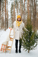 Portrait of a charming young woman holding wooden sleigh and fir tree in a snowy winter forest. She's standing on a long path between trees.