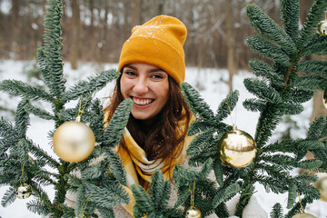 Happy smiling girl outside at winter behind synthetic fir tree decorated with shiny christmas...