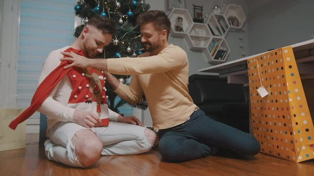 Happy homosexual couple unpacking the gifts. Man giving christmas ugly sweater to his partner. High quality 4k footage