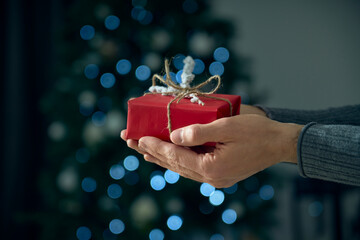 Close up shot of male hands holding a red and white gift wrapped with twine. The focus is on the box, on bokeh effect background. A present to Christmas, birthday, Valentine's Day and anniversary.