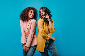 Charming african young woman looking at friend. Indoor portrait of two happy girls standing on blue...