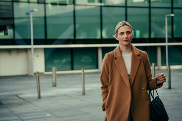 Caucasian blonde business woman looking elegant while standing in front of office building holing handbag