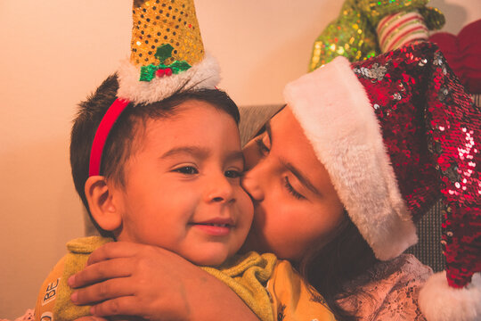 Niño y niña con gorros navideños. Hermana besa a su hermanito en el sofá de su casa.