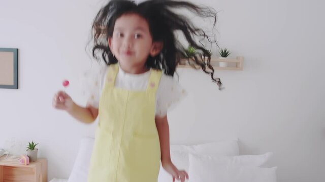 Asian little girl in a yellow dress holding lollipop and jumping for fun and relax on the bed on holiday day - Powered by Adobe