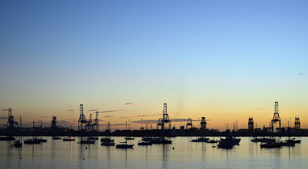 Backlit port at dawn with anchored sailboats, large cranes and totally clear blue sky