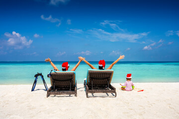 A happy family wearing santa claus hats sits in sunbeds on a tropical beach and enjoys their...