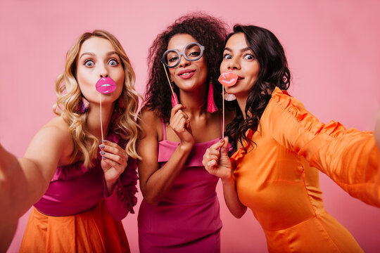 Elegant ladies making faces on pink background. International friends having fun together.