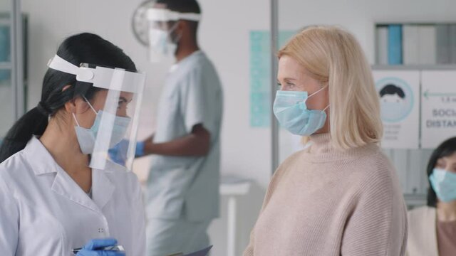 Asian female doctor in protective face screen and mask giving consultation to blonde mid-aged woman in medical office during coronavirus pandemic