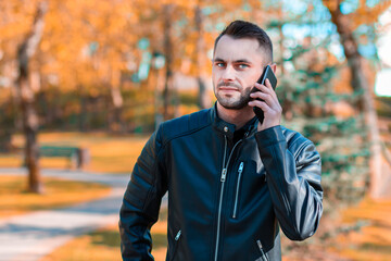 Handsome Young Man Talking on the Phone at the Beautiful Autumn Park. Youthful Guy Using Smartphone for the Call Outdoors at Sunny Day