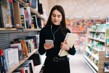 Young woman in earphones using smartphone and holding book in hand