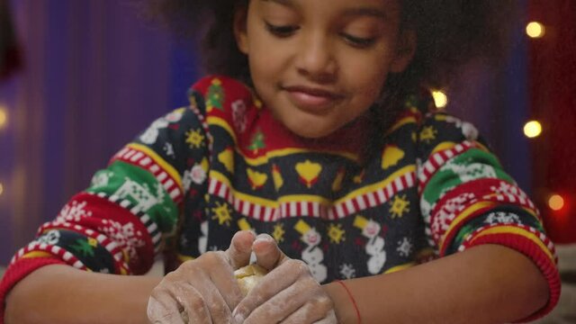 Little African American girl in a Christmas print sweater prepares dough for homemade cookies and has fun clapping her hands. Happy holidays concept. Slow motion. Close up.