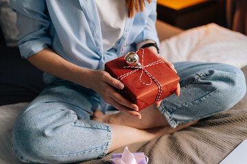 Fototapeta premium Close-up hands of unrecognizable woman holding gift box with holiday present during online communication using laptop. Happy lady sitting on lotus position at bed at bedroom and having video chat.