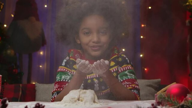 Little African American girl in New Years print sweater has fun, blows flour off her hands and prepares dough for homemade cookies. Happy holidays concept. Slow motion. Close up.