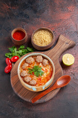 Vertical view of tomato meatballs soup with noodles in a brown bowl and different spices on dark background