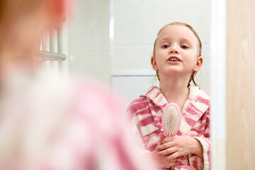 Smiling little kid girl wearing a robe looking to mirror and singing to hair brush or comb at home bathroom