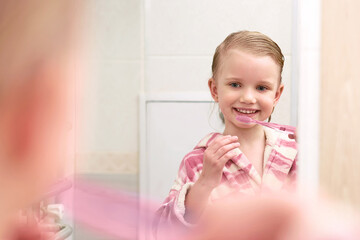 Smiling little kid girl wearing a robe looking to mirror with toothbrush, brushes teeth and smiles at home bathroom