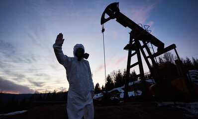 Scientist showing stop sign while standing on territory of oil field with pump jack, wearing...
