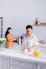 Hispanic lesbian woman holding coffeemaker near orange juice and partner on blurred background