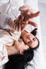 Top view of Hispanic lesbian couple touching hands on blurred foreground while lying on bed at home