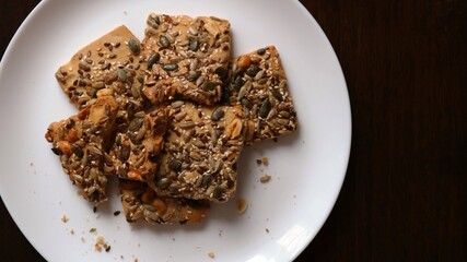 crispy crumbly cookies with peanuts and flax, sesame, sunflower and pumpkin seeds on a large white dish visible from a high angle against a dark brown background, homemade sweets on a food backdrop