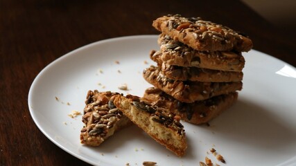 a stack of square cookies sprinkled with nuts and seeds, with one broken, on a large white plate on a dark wooden table, a homemade treat, or a sweet, healthy snack with vegan ingredients