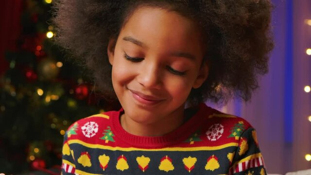 Cute curly haired African American girl enjoys trying the icing to decorate the cookies. Concept of merry new year holidays and warm family evenings. Close up. Slow motion.