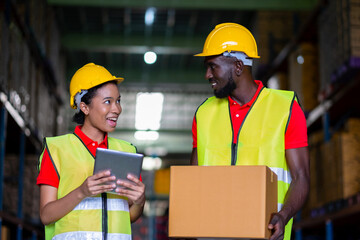 Woman worker using a tablet to check stock online and a man holding a parcel in the automotive parts  warehouse center. Both engineers people wear a safety helmet. In background shelves with goods