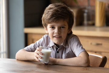 I need milk to grow healthy. Portrait of cute smiling junior boy sitting by kitchen table looking at camera holding drinking glass of dairy product. Eco farm cultured yogurt. Fermented natural kefir