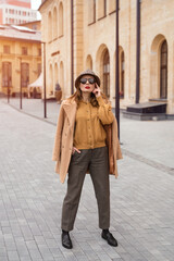 Fototapeta na wymiar Full height self confident girl in an autumn beige coat and sunglasses, plaid panama hat standing posing on the street with coat on her shoulders. Retro toned photo. 