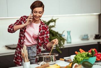 Young housewife talking on the phone while mixing a sup in a pan with a short hairstyle while prepares lunch for family in the kitchen. Healthy food at home. Horizontal image. 