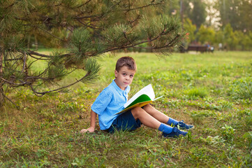 Little boy a schoolboy in a park at sunset reads a book. A surprised joyful child in a blue shirt with a yellow book in the park sits on the grass at sunset. Distance learning has ended.