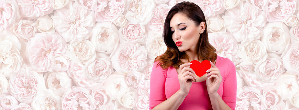 Valentine's day. A pretty Caucasian girl is holding a red paper heart.The background is a wall of pink flowers. copy space.