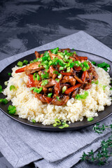 Stir fried turkey pieces with mushroom, carrot and garlic on top of couscous. Appetizing hot meal garnished with green onion served on the grey table