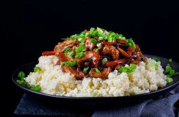 Stir-fried turkey pieces with mushroom, carrot and garlic on top of couscous on the black plate. Appetizing hot meal garnished with green onion against the black background