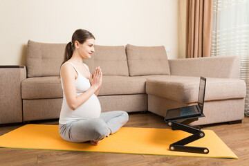 Young happy healthy yoga fitness beautiful pregnant woman doing bakasana yoga workout on yoga mat opposite laptop with online master class. lifestyle concept at coronavirus time