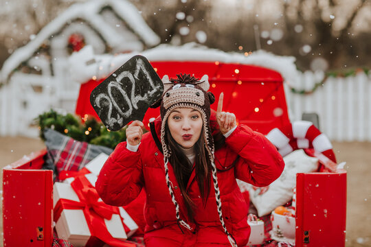 Young pretty woman in red winter jacket and knitted hat like a bull posing with name plate 2021 in the open red car with Christmas decor. Snowing.