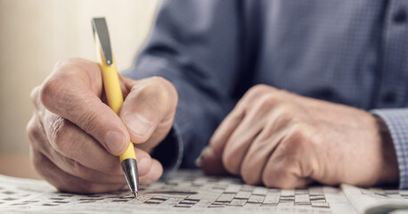 Aduld man with wrinkled skin writing letters with right hand in crossword in newspaper