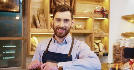 Close up of handsome young joyful Caucasian man seller in apron standing in bakery shop and putting...