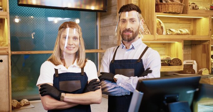 Portrait of Caucasian happy young married couple bakers in protective face shields and gloves holding basket with fresh bread standing in small own bakehouse in quarantine. Family business concept