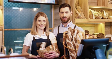 Close up of joyful happy Caucasian young married couple in good mood holding baskets with fresh bread standing in small own bakery shop and smiling to camera. Man and woman seller in bakehouse