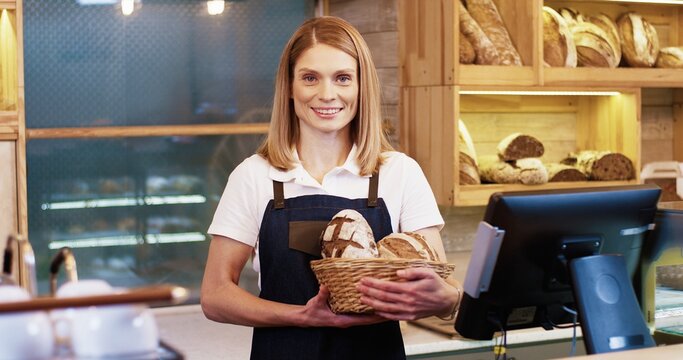 Portrait of cheerful beautiful young Caucasian woman seller bakery owner in apron standing in small store and holding basket with fresh baked bread in hands, looking at camera and smiling alone