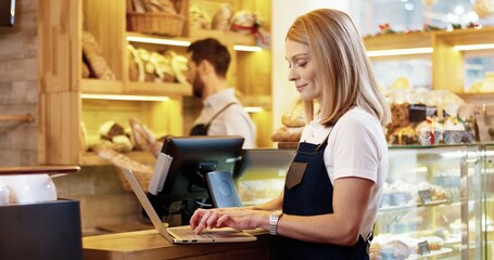 Close up of beautiful young Caucasian woman seller in apron standing in bakery, tapping and typing on laptop talking to male colleague working on background. Bakehouse concept. Small business