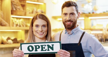 Close up of cheerful Caucasian young male and female bakers standing in own bakery and holding Open...