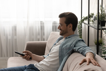 Good time by tv set. Side view of relaxed young man sitting on sofa watching tv, choosing cable...