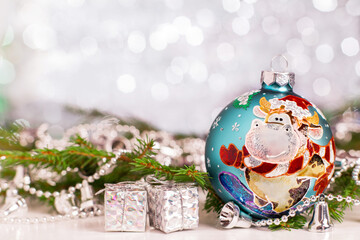 Christmas decoration - a hand-painted ball with the symbol of the Year of the Ox on a snowboard. Nearby is a tree branch and New Year's decorations. Blurred background of silver color