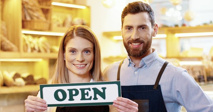Close up of cheerful Caucasian young male and female bakers standing in own bakery and holding Open sign in hands, looking at camera and smiling. Bakehouse reopening. Small business opening - Powered by Adobe